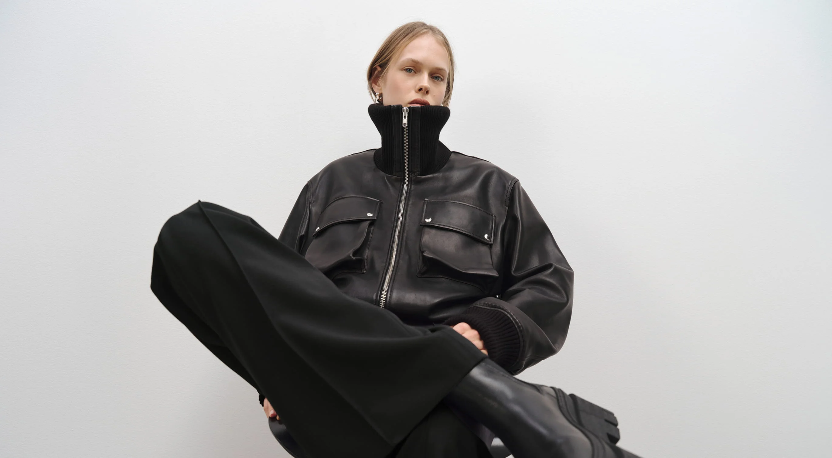 Model in schwarzer Lederjacke und weiter schwarzer Hose sitzt vor einer weißen Wand.